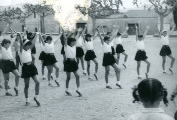 Schwarzes und weißes Bild von jungen Mädchen in Schuluniformen, die auf einer Straße tanzen, die von Gebäuden und Bäumen gesäumt ist, mit einer Person in Schuhen am unteren Rand.