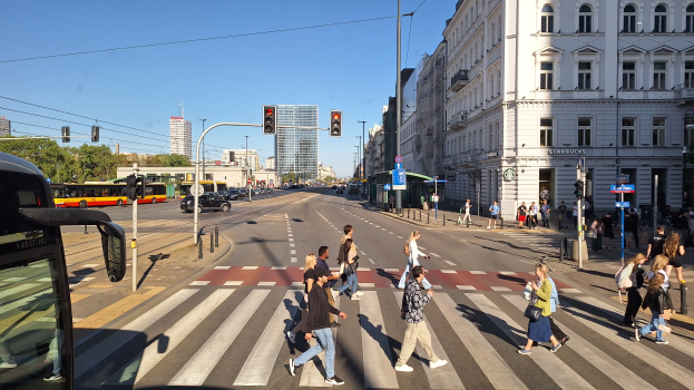 Gruppe von Menschen, die eine Straße an einer Ampel überqueren, mit einem Bus auf der linken Seite, Fahrzeugen auf der Straße, Verkehrszeichen, Laternenmasten, Gebäuden, Bäumen und einem klaren blauen Himmel.