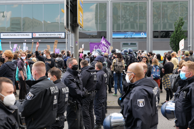 Eine große Gruppe von Menschen demonstriert vor einem Gebäude, einige halten Schilder und tragen Helme, mit einem Schildständer und einem Baum im Vorder- und Hintergrund.