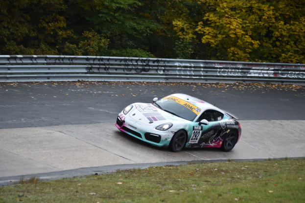 Ein Porsche Cayman GT4 auf der Nürburgring 24 Stunden Rennstrecke, umgeben von grünem Gras und Bäumen, mit Text und Zahlen auf dem Auto und einem Geländer entlang der Strecke.