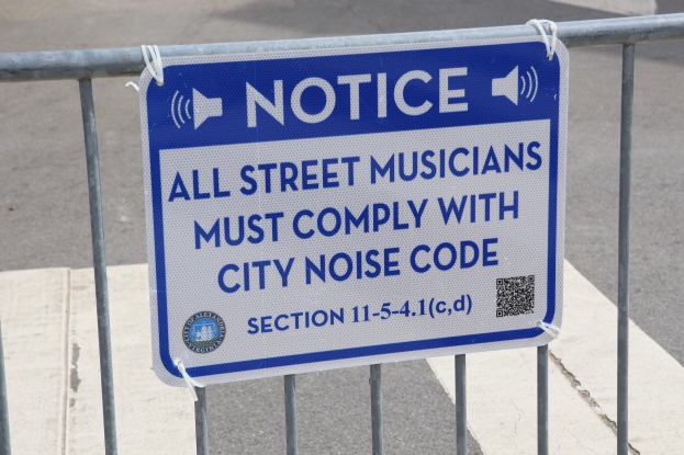 Anschlagtafel an einem Zaun mit einem Schild, das Straßenmusikern den Einhaltung des städtischen Lärmschutzes vorschreibt, mit einer Straße im Hintergrund.
