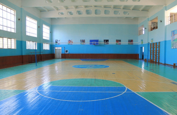 Großer Turnhallen mit einem blauen und weißen Boden, ausgestattet mit einem Basketballfeld, Netz, Stangen, einer Tür, Fenstern, Wandplakaten, einem Geländer und einem Dach.