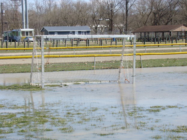 Ein Fußballtor steht in der Mitte eines überfluteten Feldes mit Gras und Wasser, umgeben von Hütten, Pfählen, Bäumen, Fahrzeugen und einem klaren blauen Himmel im Hintergrund.