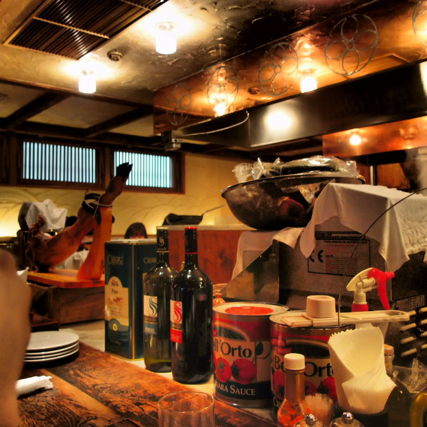 Ein unordentlicher Raum mit einem Tisch, der mit Weinflaschen, Tellern, Schachteln, Taschentüchern und Papptellern gefüllt ist, und zwei Personen in der Nähe offener Fenster sitzen.