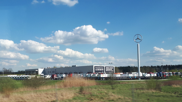 Ein Mercedes-Benz-Werk in Deutschland mit Fahrzeugen auf der Straße, umgeben von Gras, Pflanzen, Bäumen, Gebäuden und Strommasten, einschließlich eines mit einem Logo; bewölkter Himmel im Hintergrund.