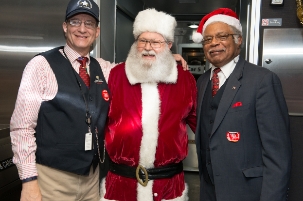 Eine Gruppe von Menschen mit Brillen, Mützen, Krawatten, Ausweisen und Abzeichen, darunter eine Person in einem Weihnachtskostüm, die vor einer Stahlwand mit Lichtern, Schildern und anderen Gegenständen im Hintergrund steht.