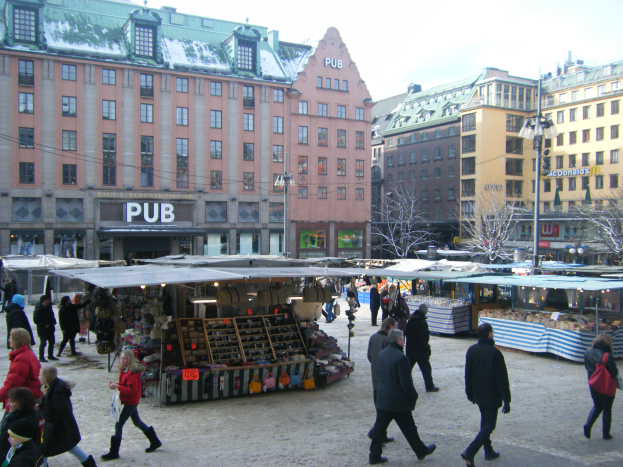 Ein lebendiger Weihnachtsmarkt in Stockholm, Schweden, mit Menschen, die herumlaufen, Ständen voller Gegenstände, Gebäuden, Laternen, Bäumen und einem klaren blauen Himmel im Hintergrund.