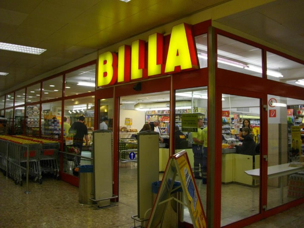 Ein Markt mit einem Eingang in der Mitte, Mülltonnen daneben, eine Reihe von Einkaufswagen auf der linken Seite und einige Menschen drinnen, die unter Deckenbeleuchtung Produkte durchstöbern.
