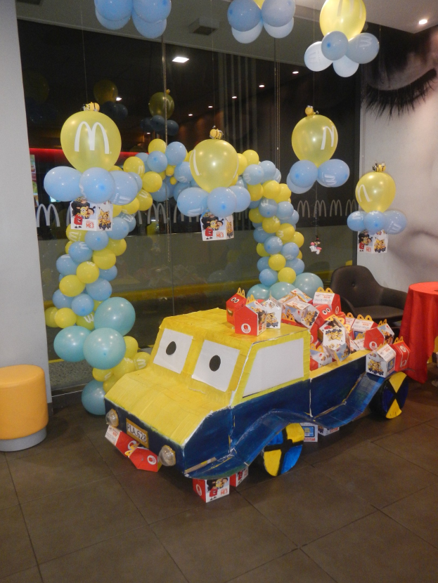 Eine festliche McDonald's-Geburtstagsfeier-Szene mit einem gelben und blauen Lastwagen, der mit Ballons und anderen Dekorationen geschmückt ist, einem Tisch mit roter Decke und Gegenständen, Hockern, einer Glaswand und Deckenlampen.