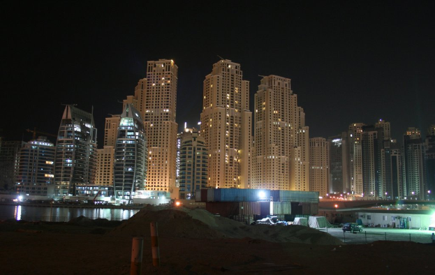 Ein schwarzes Auto steht auf sandigem Boden neben Kisten, mit Gebäuden, Straßenlaternen und Pfählen im Hintergrund, unter einem dunklen Himmel und einem Gewässer auf der linken Seite.