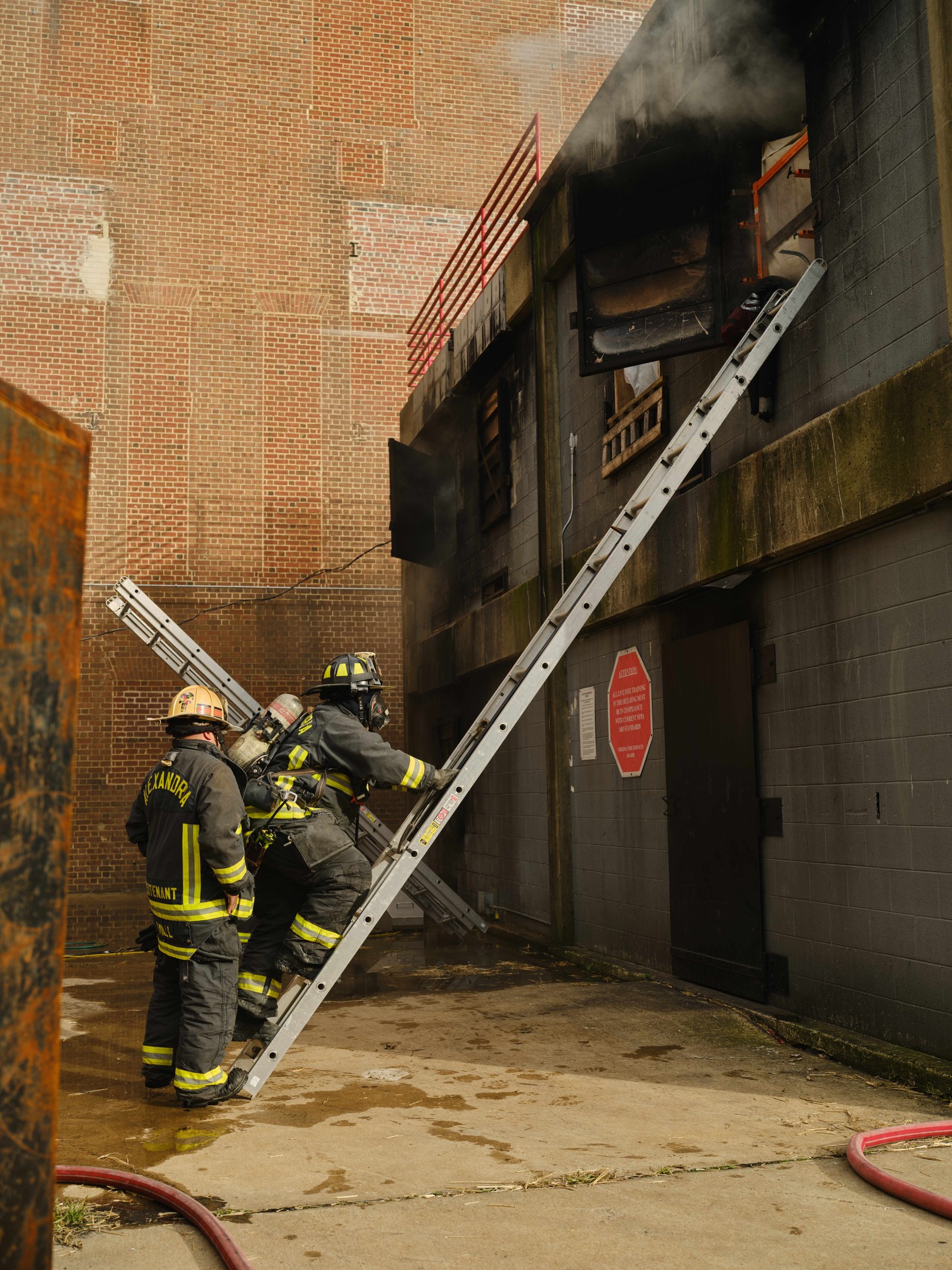Feuerwehrleute in Helmen klettern eine Leiter zu einem rauchgefüllten Gebäude hinauf, mit einem Metallobjekt links und Rohren an der Basis.