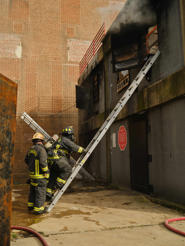 Feuerwehrleute in Helmen klettern eine Leiter zu einem rauchgefüllten Gebäude hinauf, mit einem Metallobjekt links und Rohren an der Basis.