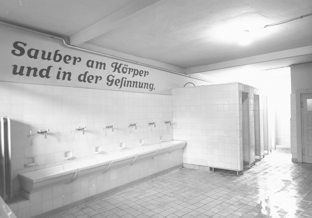 Ein Schwarz-Weiß-Foto einer öffentlichen Toilette mit einem gefliesten Boden, einer textbedeckten Wand, mehreren Hängen, einer Tür und einer Deckenleuchte.