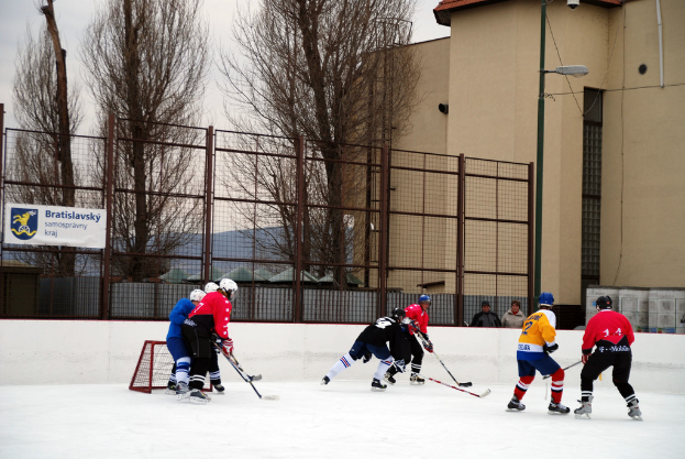 Menschen, die Eishockey auf einer Eisbahn mit Gebäuden, Bäumen, einer Straßenlaterne, einem Namensschild und Zäunen im Hintergrund unter einem Himmel spielen.
