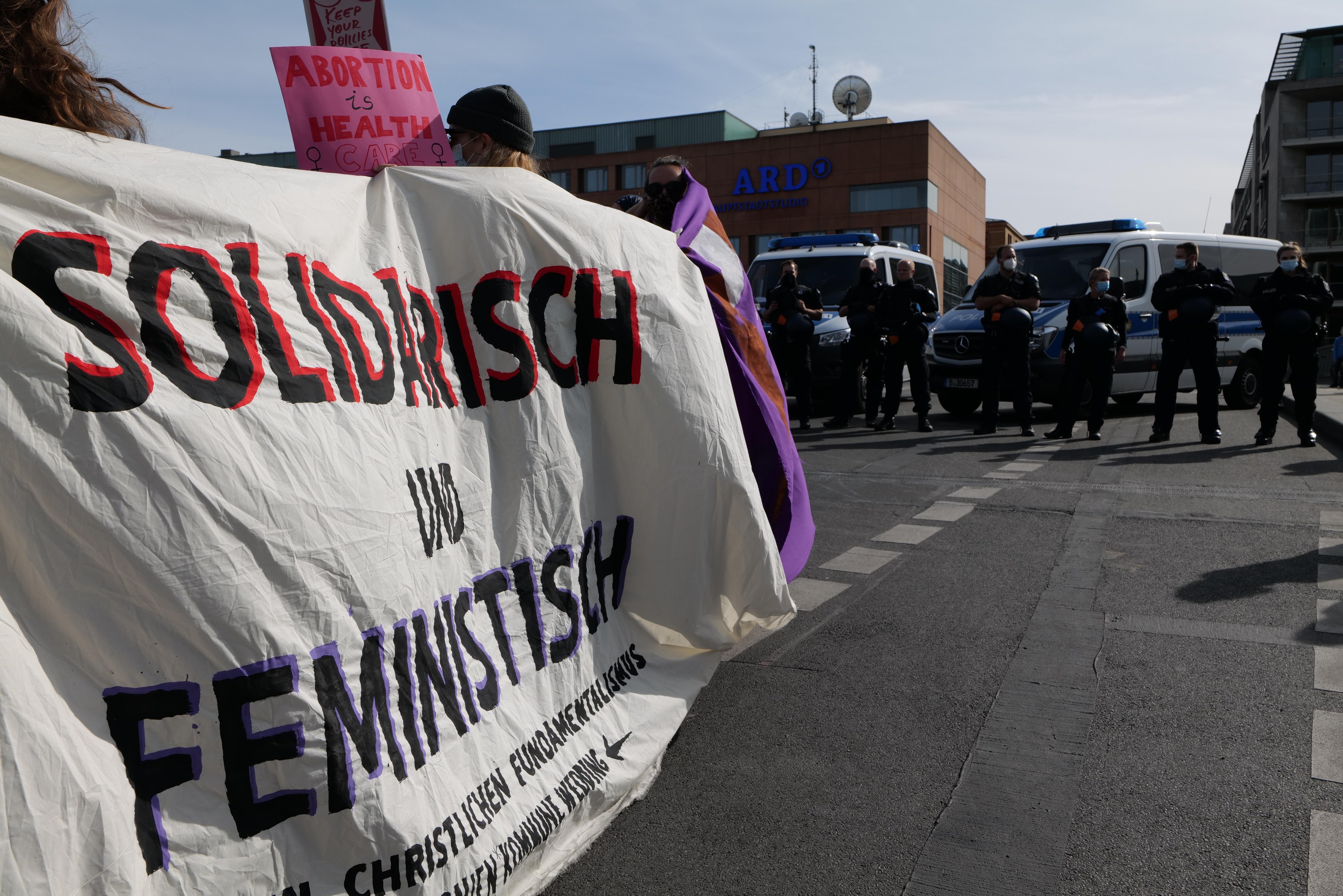 Gruppe von Menschen, die eine 'Solidarität und Feminismus'-Fahne tragen, öffentlich auf einer Straße mit geparkten Fahrzeugen und Gebäuden im Hintergrund bei klarem blauem Himmel.