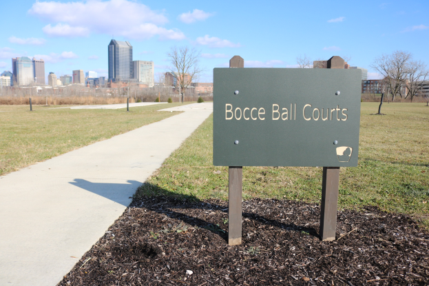 Ein Schild am Boden neben einem Weg und Bäumen trägt die Aufschrift "Bocciaball-Plätze in Indianapolis, Indiana" mit einem bewölkten Himmel darüber.