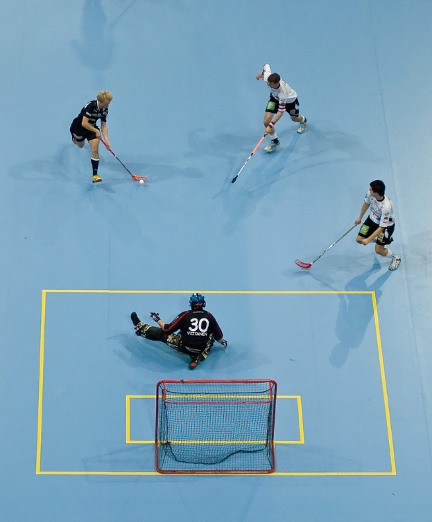 Drei Personen in Sportkleidung spielen Hockey, wobei eine Person rechts ein Ball mit einem Hockeystock schlägt und eine Person hinter einem Netz auf dem Boden sitzt.