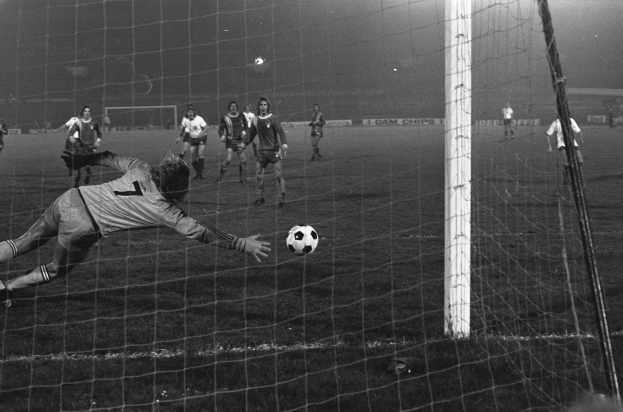Ein Schwarz-Weiß-Foto eines Fußballspielers, der vor einem Tor einen Ball hält, umgeben von einer Gruppe von Menschen auf dem Boden. Das Torpfosten ist auf der rechten Seite des Bildes zu sehen, und im Hintergrund gibt es Werbetafeln und Lichter.