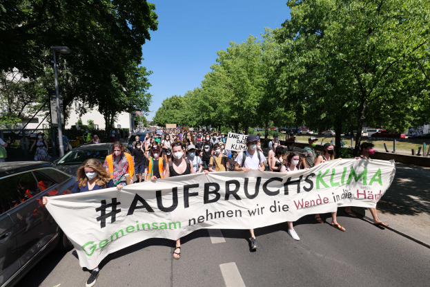 Eine Gruppe von Menschen geht eine Straße entlang und hält ein Schild hoch, auf dem 'Aufbrucksklima' steht und trägt Masken, mit Fahrzeugen, Bäumen, einem Laternenpfahl, Gebäuden und einem klaren blauen Himmel im Hintergrund.