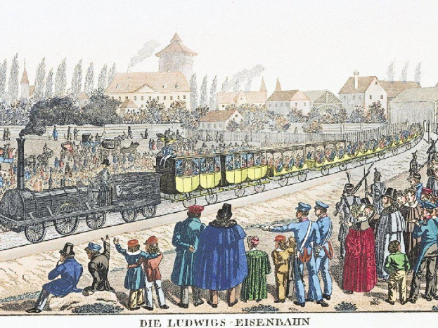 Eine Gruppe von Menschen steht um einen frühen Zug auf den Schienen mit einigen Passagieren im Inneren und anderen auf dem Boden, mit Gebäuden und Bäumen im Hintergrund und dem Text "Ludwigs-Eisenbahn, die erste Bahnstation in Deutschland" unten.