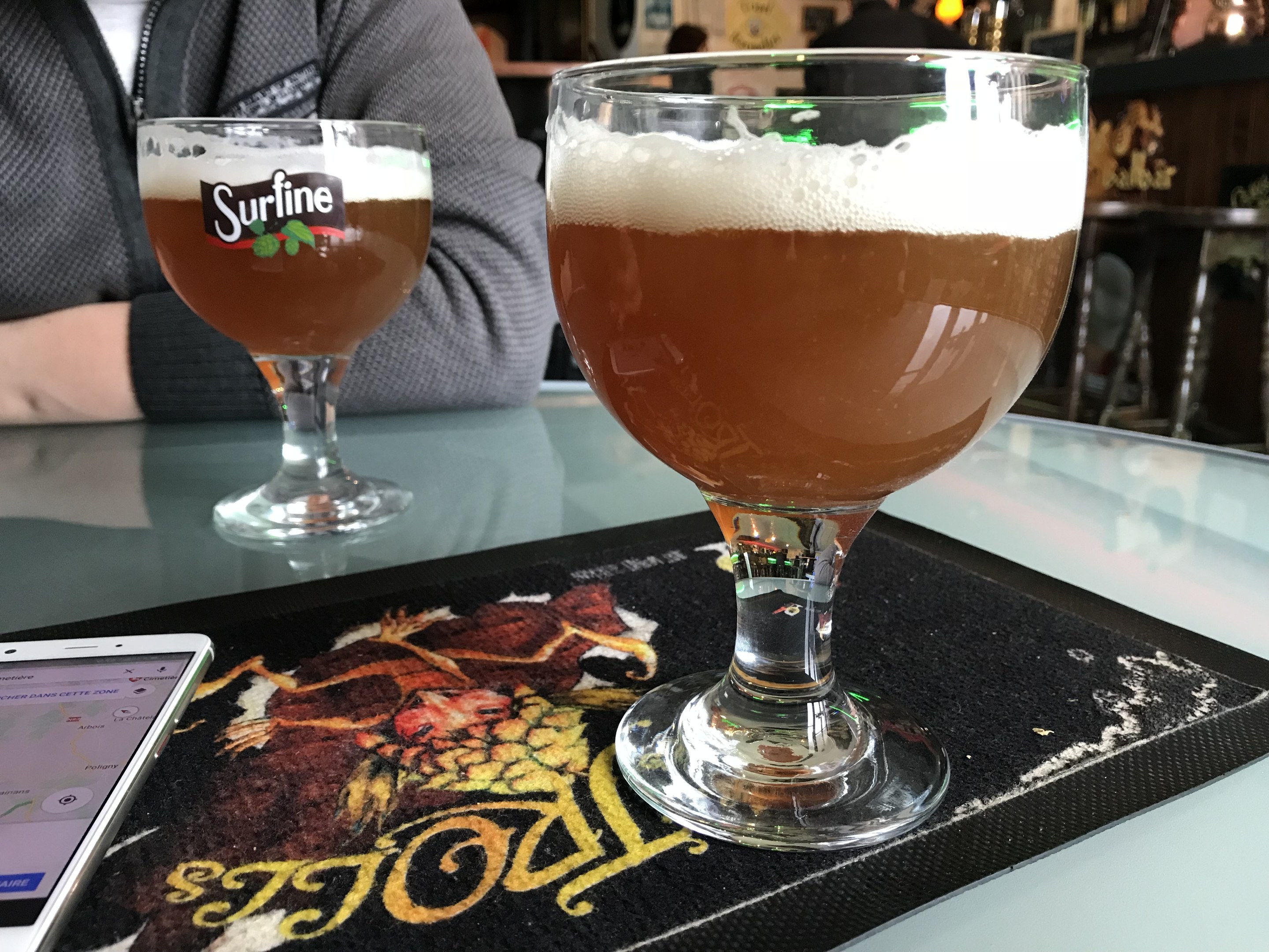 Zwei Gläser goldenen Bieres auf einem Tisch mit einem Handy in der Nähe, umgeben von Menschen und gerahmten Kunstwerken an der Wand.