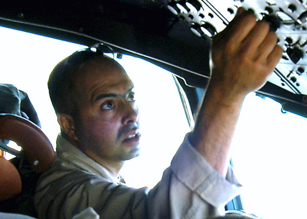 Ein Mann in einem blauen Hemd und schwarzer Mütze sitzt im Fahrersitz eines hellblauen Autos und hält das Lenkrad mit entschlossener Miene, wobei er wie ein Taxifahrer mit getönten Fenstern wirkt.