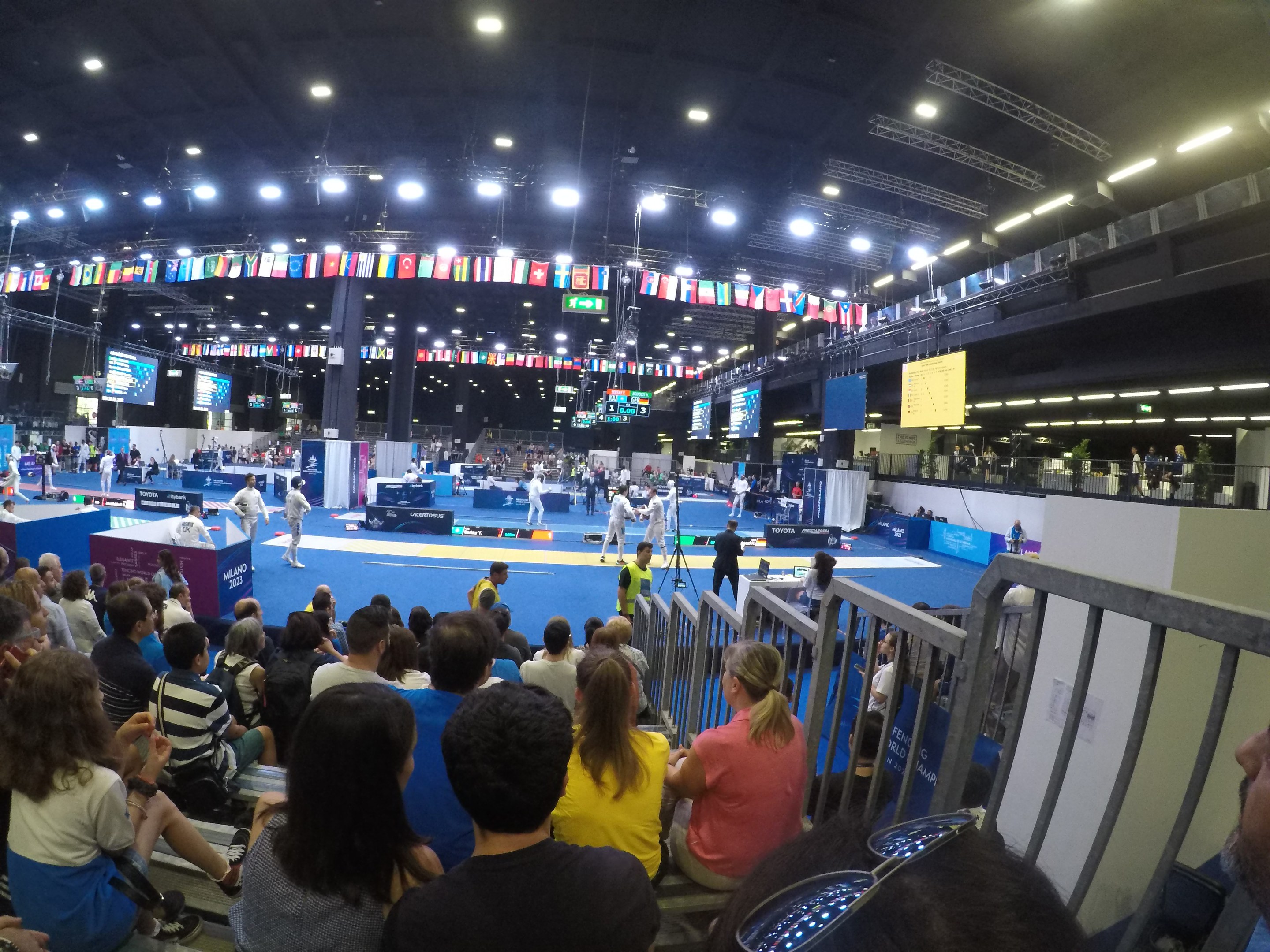 Eine große Menschenmenge auf Sitzplätzen und Stehplätzen in einer Indoor-Sportarena mit Bildschirmen, Fahnen und Deckenleuchten im Hintergrund.