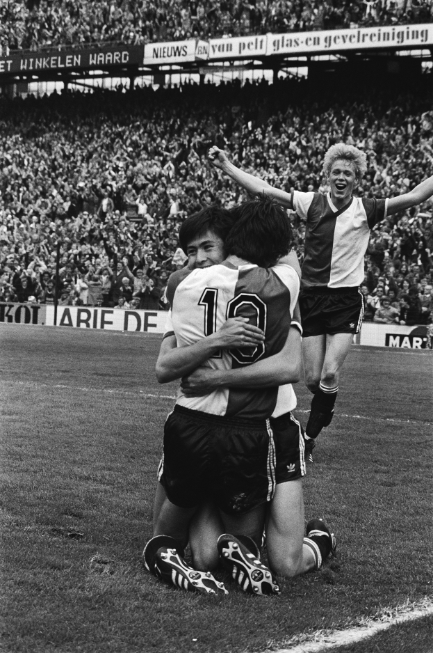 Drei Fußballspieler feiern Tor auf dem Boden vor jubelnder Menge in einem schwarz-weißen Stadion.