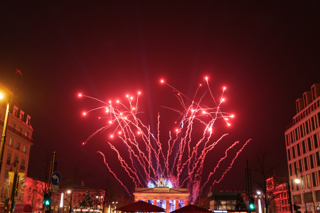 Eine Stadtstraße in Berlin an Silvester, voller Gebäude, Bäume, Laternenpfähle, Verkehrszeichen, Zelte und Menschen, mit einem von Feuerwerk erleuchteten Himmel im Hintergrund.