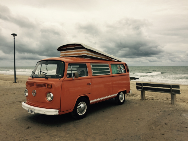 Ein oranger VW-Bus auf einem sandigen Strand neben dem Ozean geparkt, mit einer Bank und einem Laternenmast in der N├Ąhe unter einem bew├Âlktem Himmel.
