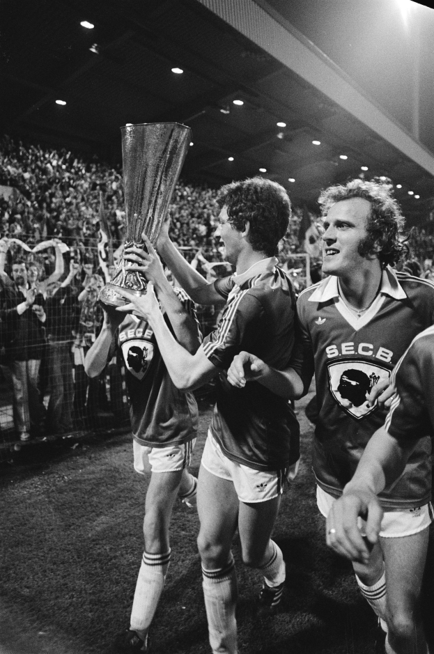 Schwarzes und weißes Foto von Fußballspielern, die mit einem Pokal auf einem Feld feiern und von einer Menge hinter einem Zaun und Stadionbeleuchtung umgeben sind.