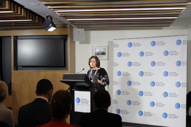 Eine Frau steht auf einer Bühne und spricht in ein Mikrofon, vor ihr sitzen Menschen auf Stühlen, an der Wand hinter ihr hängt ein Banner, auf dem Holzwand steht ein Fernseher und an der Decke leuchten Lampen.