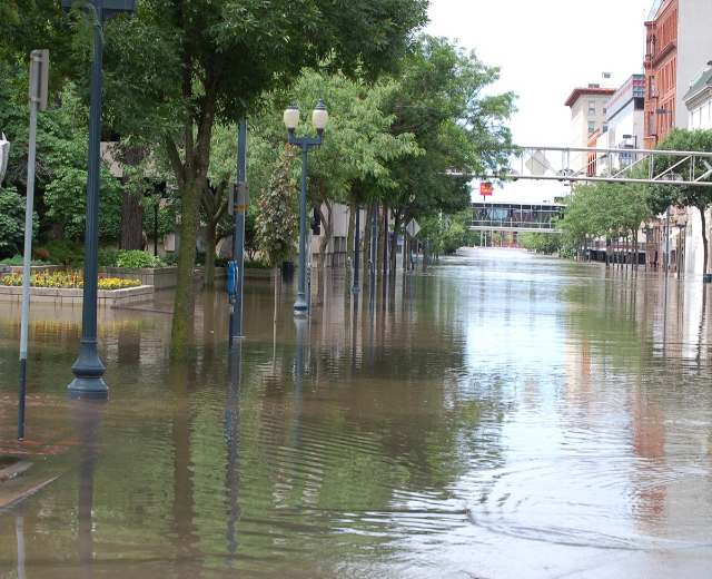Flutstraße in der Stadtmitte mit Wasser, das die Straße, die Straßenmöbel, die Vegetation und die benachbarten Gebäude bedeckt, und eine Brücke im Hintergrund unter einem bewölkten Himmel.