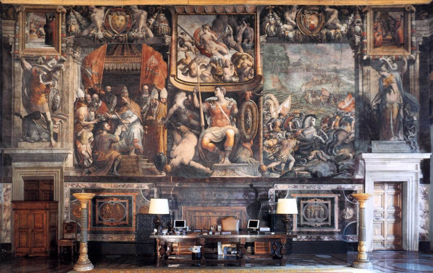 Großer Raum mit einem prominenten Gemälde an der Wand, einem Tisch mit Gegenständen, zwei Lampen, zwei Türen und einem Teppich, befindet sich im Palazzo della Signoria in Florenz, Italien.