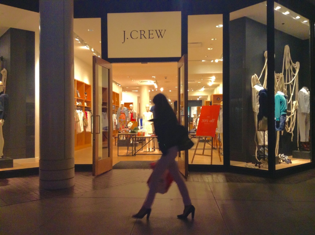 Ein Einkaufszentrum mit Glaswänden und -türen, das Schaufensterpuppen, Tische, hängende Kleider und Schränke zeigt, mit einer Frau daneben.