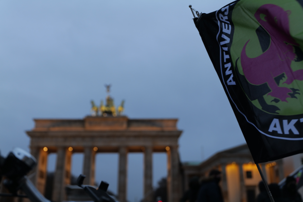 Eine Gruppe von Menschen steht vor dem Brandenburger Tor in Berlin, Deutschland, mit einem Fahnenmast mit Logo und Text im Vordergrund, die sich versammelt haben, um gegen die antifaschistische Bewegung zu protestieren.