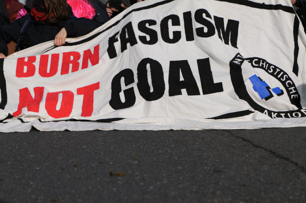Eine Gruppe von Menschen steht auf einer Straße und hält ein buntes Banner mit der Aufschrift 'Verbrenne Faschismus nicht Ziel'.