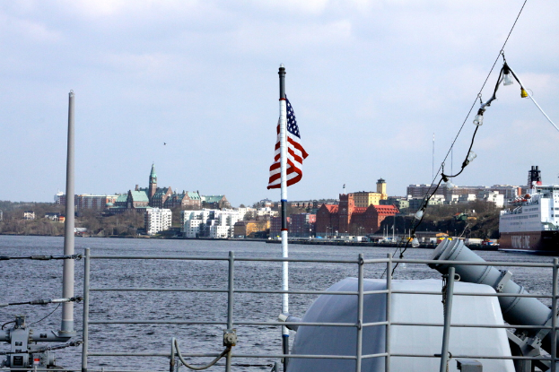 Eine Stadtansicht mit einem Schiff im Vordergrund, das eine Flagge zeigt, Booten auf dem Wasser, Gebäuden, Bäumen und Pfählen im Hintergrund, unter einem bewölkten Himmel.