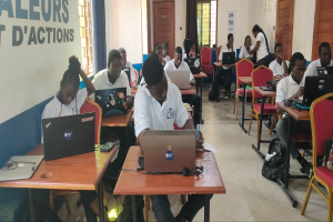 Eine Gruppe von Menschen sitzt an Tischen mit Laptops in einem Klassenzimmer, mit Taschen auf dem Boden und Text an der linken Wand, was auf einen Online-Kurs in Ghana hindeutet.