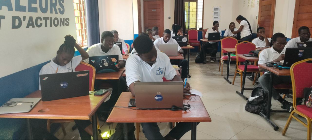 Eine Gruppe von Menschen sitzt an Tischen mit Laptops in einem Klassenzimmer, mit Taschen auf dem Boden und Text an der linken Wand, was auf einen Online-Kurs in Ghana hindeutet.