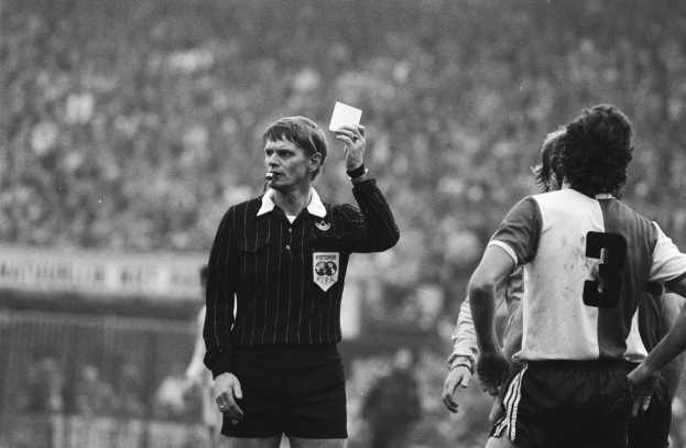 Ein Schiedsrichter in einem Schwarz-Weiß-Bild hält eine rote Karte vor einer Menge von Menschen in Fußballuniformen, mit einem Zaun im Hintergrund.