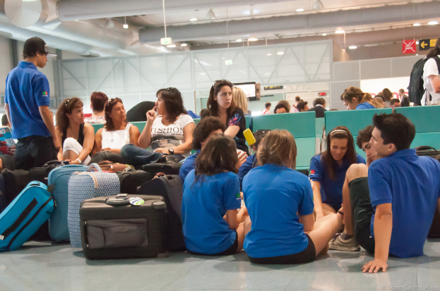 Ein belebter Flughafeninnenraum mit vielen auf Fl├╝ge wartenden Menschen, Gep├Ąck links und Glast├╝ren mit Hinweisen im Hintergrund.