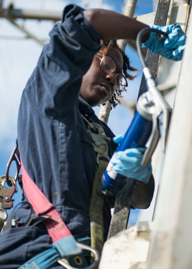 Mann in blauem Hemd, blauen Handschuhen und Brille arbeitet an einer Stromleitung und hält eine Bohrmaschine, mit Metallstangen im Hintergrund.