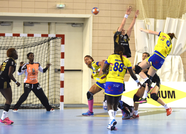 Eine Gruppe von Frauen beim Handballspielen auf einem Platz mit einem Ball in der Luft, ein Tor mit Netz, eine Fahne mit Text, ein Vorhang und eine Wand im Hintergrund.