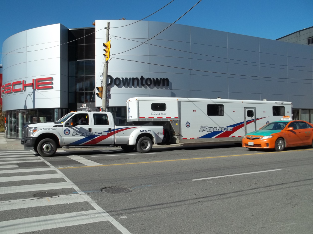 Ein Polizeiwagen vor einem Gebäude mit Text, eine Ampel mit einem Mast und Drähten im Vordergrund und Bäume mit einem klaren blauen Himmel im Hintergrund.