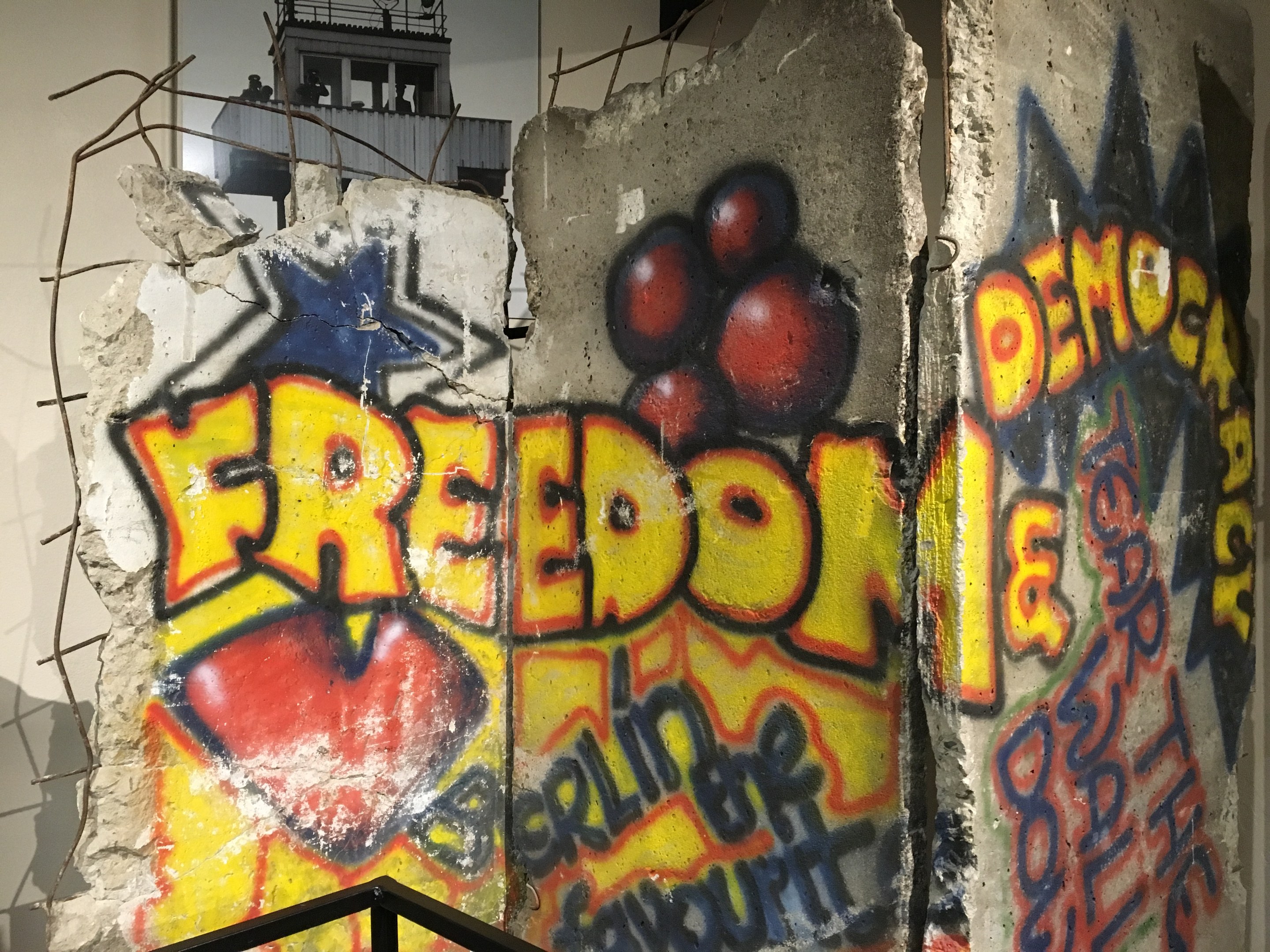 Eine Wand mit Graffiti, das "Freiheit und Demokratie" in großen schwarzen Buchstaben lautet, ein Geländer unten und ein Gebäude mit Fenstern im Hintergrund.