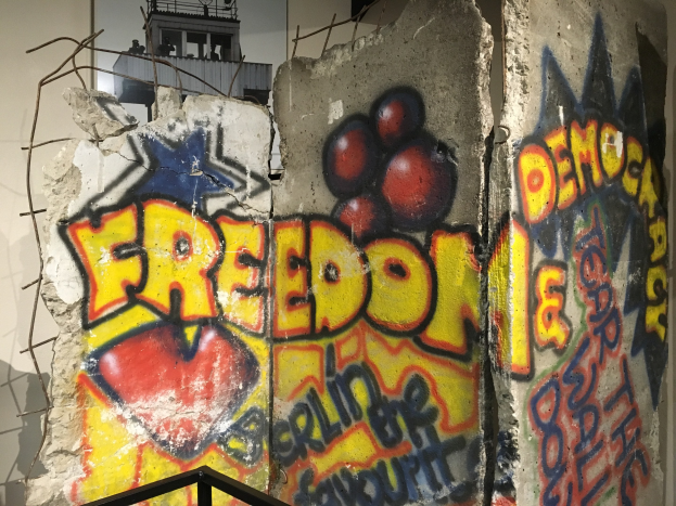 Eine Wand mit Graffiti, das "Freiheit und Demokratie" in großen schwarzen Buchstaben lautet, ein Geländer unten und ein Gebäude mit Fenstern im Hintergrund.