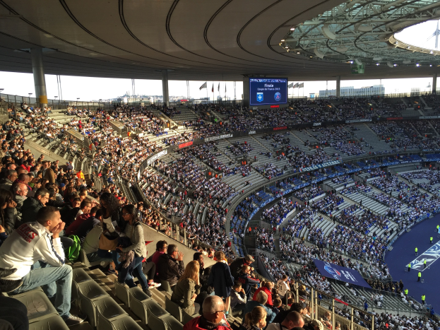 Große Menschenmenge in einem Stadion bei einem Förballett, mit einer Bühne auf der rechten Seite, Fahnen, Masten, einem Bildschirm und der Allianz Arena in München, Deutschland im Hintergrund.