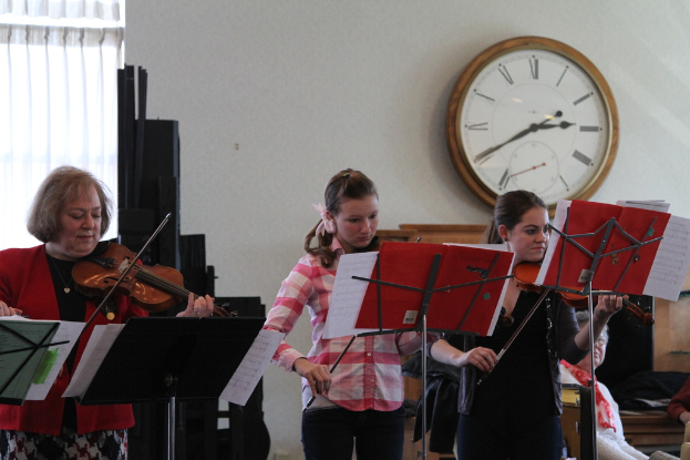 Drei Frauen auf der Bühne mit Geigen spielend, mit Notenpulten und Noten vor ihnen, während das Publikum im Vordergrund sitzt und eine Uhr an der weißen Wand im Hintergrund hängt, mit einem weißen Vorhang in der linken Ecke.