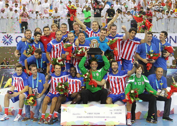 Eine Gruppe von Menschen in Sportkleidung, einige halten Auszeichnungen und Blumen, steht vor einer Menge mit Geländern und Schildern im Hintergrund.